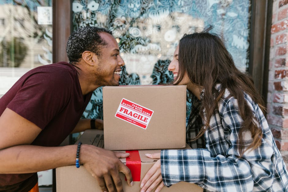 Two women stand inside a room surrounded by packed cardboard boxes, some sealed with red tape and labeled, indicating they are preparing for a house move. One woman, with light skin and wearing a chec