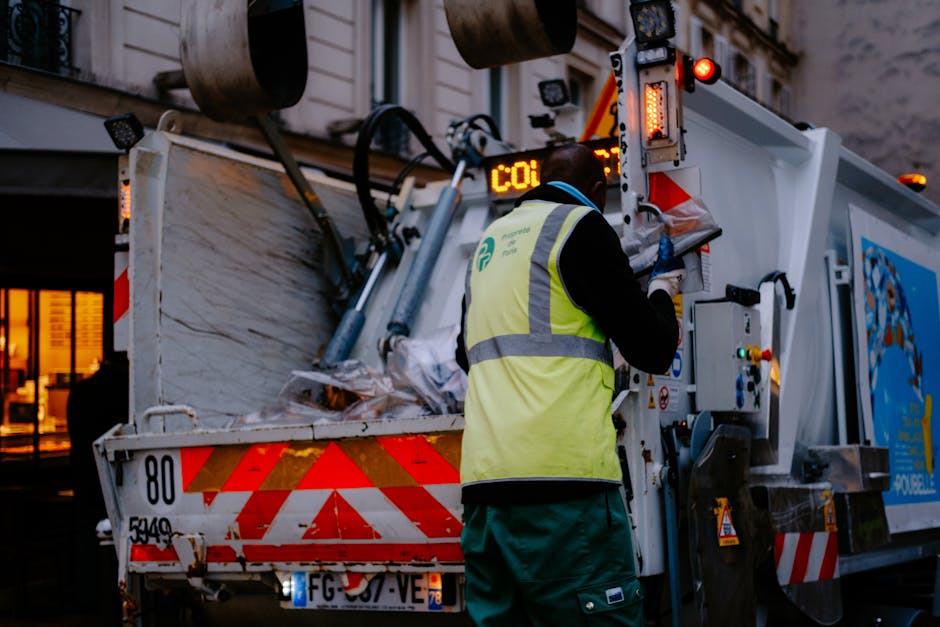 A worker wearing a high-visibility yellow vest and blue gloves is operating a waste collection vehicle on a street during dusk or early evening. The vehicle, branded with a blue and white logo, has its rear open, revealing a large compartment filled with various types of rubbish, including cardboard boxes, plastic packaging, and mixed waste. The worker is seen using controls mounted on the side of the vehicle, possibly to operate the waste compaction or lifting mechanisms. Nearby, a building with lit windows is visible, indicating an urban residential or commercial area. The scene emphasizes waste removal or clearance processes associated with house or office relocations, consistent with the services offered by Man With a Van Wallington, especially related to bulk waste removals and moving logistics.