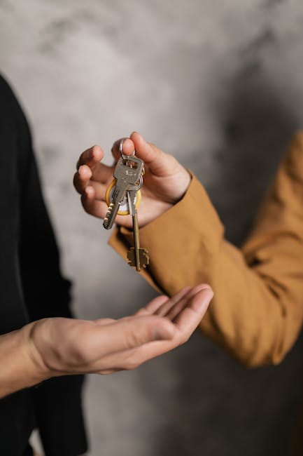 A person dressed in a black outer garment is handing over a set of brass keys to another individual wearing a brown jacket. The background features a neutral grey wall with a textured surface, suggesting an indoor setting, possibly an entrance or corridor related to a home or property. The keys, which include a standard house key and a smaller auxiliary key, are held firmly in the hand of the recipient, who extends their palm to receive them. This exchange occurs within a context that could be associated with home relocation or handover procedures during moving or property transfer. The lighting is soft and evenly distributed, highlighting the keys and the hands involved in the transaction. As part of the moving process, [COMPANY_NAME] often assists with key handovers during furniture transport or home removals, ensuring a smooth transition from previous to new occupants.