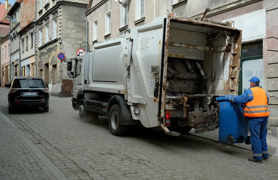 A worker wearing a high-visibility yellow vest and blue gloves is operating a waste collection vehicle on a street during dusk or early evening. The vehicle, branded with a blue and white logo, has its rear open, revealing a large compartment filled with various types of rubbish, including cardboard boxes, plastic packaging, and mixed waste. The worker is seen using controls mounted on the side of the vehicle, possibly to operate the waste compaction or lifting mechanisms. Nearby, a building with lit windows is visible, indicating an urban residential or commercial area. The scene emphasizes waste removal or clearance processes associated with house or office relocations, consistent with the services offered by Man With a Van Wallington, especially related to bulk waste removals and moving logistics.