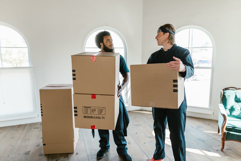 Two movers from Man With a Van Wallington are inside a well-lit residential room with light-colored wooden flooring and large arched windows allowing natural daylight. One mover, with curly hair and a beard, is holding a large cardboard box with red tape, while the other, wearing a bandana, is gesturing towards him and carrying a similar box. There are additional cardboard boxes stacked on the floor nearby, some plain and others labeled with dimensions, indicating the packing process involved in home relocation. The room contains minimal furniture, including a green upholstered armchair with wooden arms on the right side, and the movers appear to be preparing the space for furniture transport and logistics of packing and moving. This scene reflects the careful handling and loading stages typical of professional removals services, highlighting the logistics of internal unpacking and packing within a house removal process.