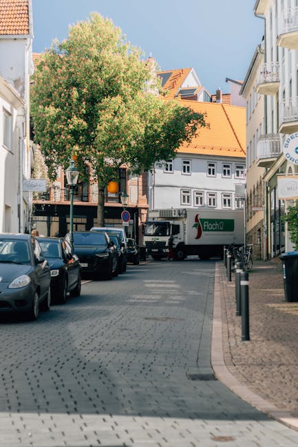 A narrow residential street in Wallington with parked cars lining both sides, including a small black hatchback and a larger van. A large green-leafed tree with dense foliage is situated on the sidewalk near the center of the image, providing partial shade to the area. The buildings on either side of the street are multi-storey, with white and beige facades, and some feature balconies and shop fronts. In the background, a delivery truck with the logo 'Flecha' is partially visible, positioned across the street near a building entrance. The street is paved with small gray cobblestones, and black bollards line the sidewalk to prevent vehicle intrusion. The lighting suggests a sunny day with clear skies, and the scene captures an area where home relocation and furniture transport could be taking place, possibly involving [COMPANY_NAME] for moving services or house removals.
