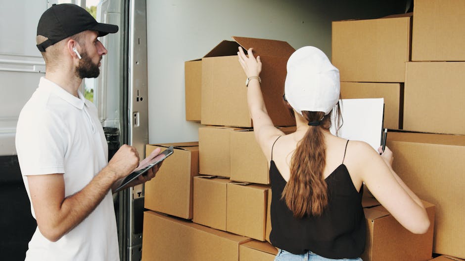 Two movers from Man With a Van Wallington are inside a well-lit residential room with light-colored wooden flooring and large arched windows allowing natural daylight. One mover, with curly hair and a beard, is holding a large cardboard box with red tape, while the other, wearing a bandana, is gesturing towards him and carrying a similar box. There are additional cardboard boxes stacked on the floor nearby, some plain and others labeled with dimensions, indicating the packing process involved in home relocation. The room contains minimal furniture, including a green upholstered armchair with wooden arms on the right side, and the movers appear to be preparing the space for furniture transport and logistics of packing and moving. This scene reflects the careful handling and loading stages typical of professional removals services, highlighting the logistics of internal unpacking and packing within a house removal process.