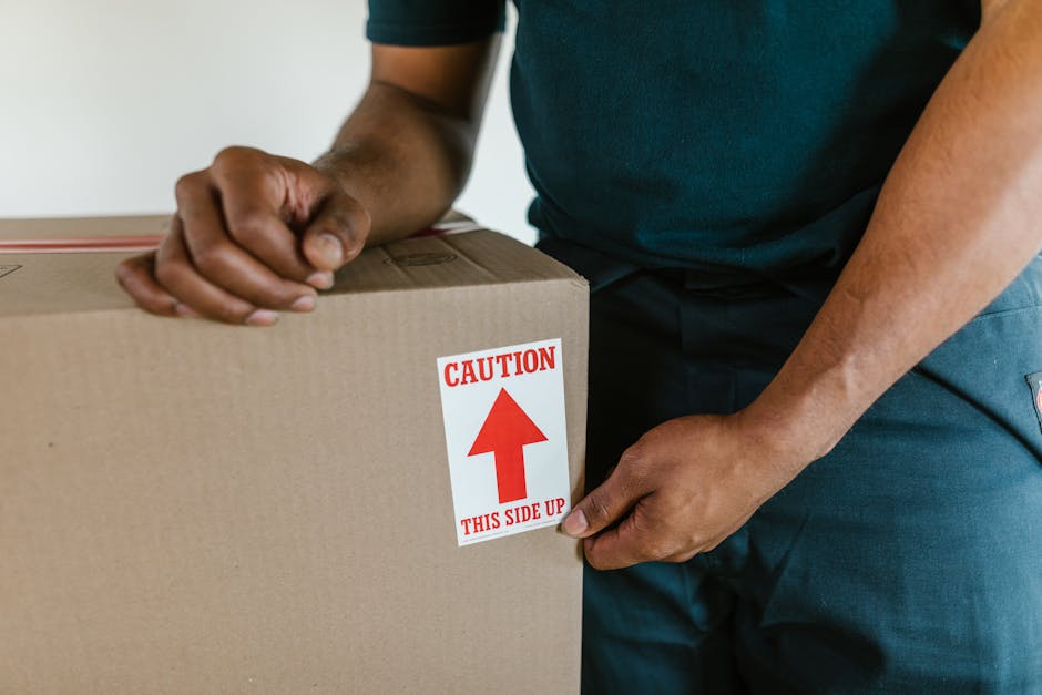Close-up of a mover's hands securing a large cardboard box with packing tape, featuring a red and white caution sticker indicating 'This Side Up,' inside a home. The mover is wearing dark clothing and is positioned near the doorway, preparing the box for transport during a house removal or home relocation. The setting suggests the packing and loading process typical for furniture transport and moving logistics, with the box being carefully handled to prevent damage during loading onto a van or for storage. The background is blurred, focusing on the manual handling of the box, highlighting the attention to safety and proper packing practices involved in professional removals, as provided by Man With a Van Wallington.