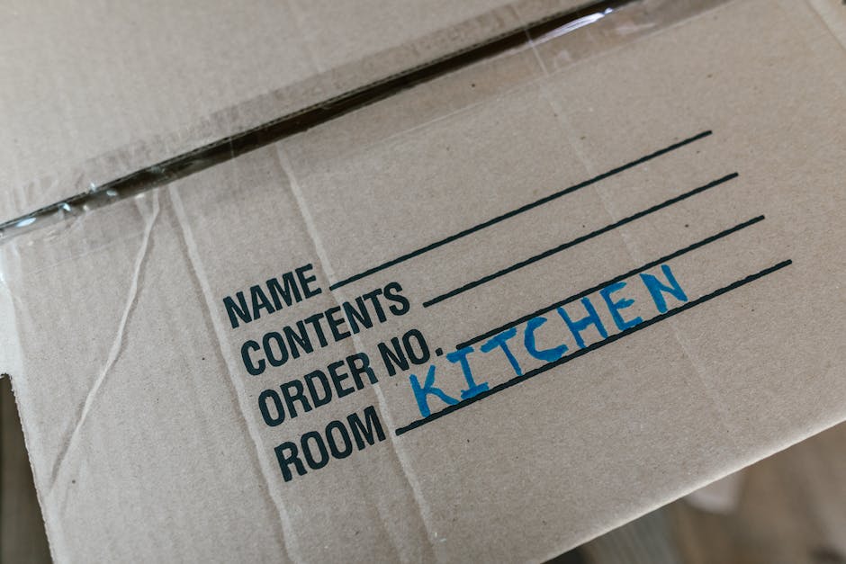 Close-up image of a cardboard moving box with printed and handwritten labels, including the words 'NAME', 'CONTENTS', 'ORDER NO.', and 'ROOM', with 'KITCHEN' handwritten in blue marker on the row designated for the room. The box appears to be sealed with clear tape and is situated on a flat surface, possibly during a home relocation or packing process. The image emphasizes organized packing and labeling, which are essential parts of furniture transport and moving services provided by Man With a Van Wallington, especially when preparing for house removals or logistics planning, ensuring items from the kitchen are properly identified for efficient loading and unloading.