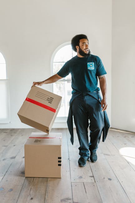 A man with dark curly hair and a beard is standing inside a bright, empty room with white walls and wooden flooring. He is wearing a blue T-shirt with a logo on the chest and dark trousers, and appears to be in the process of moving. He is holding a cardboard box wrapped with red tape in his right hand while another box is placed on the floor beside him. The room features a large arched window in the background, letting in natural light, with minimal furnishings and no visible clutter. The scene depicts packing and moving activities, possibly part of a home relocation service provided by Man With a Van Wallington. The environment suggests a professional approach to transportation and logistics related to house removals, with emphasis on careful handling of packed boxes during a furniture transport or packing and moving process.