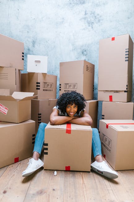 A young woman with curly black hair, wearing a white shirt, blue jeans, and white sneakers, is sitting on the wooden floor inside a room with a distressed pale blue wall. She is smiling and leaning on a large cardboard box with red packing tape, surrounded by numerous other unpacked cardboard boxes of various sizes, some closed and others partially open. The boxes are positioned against the wall and scattered across the floor, indicating a home relocation process. The room appears to be in the midst of a packing or unpacking phase, with some boxes labeled for transport. The setting suggests the early stages of furniture transport and household packing, which is relevant to the services offered by Man With a Van Wallington in the context of house removals and moving logistics.