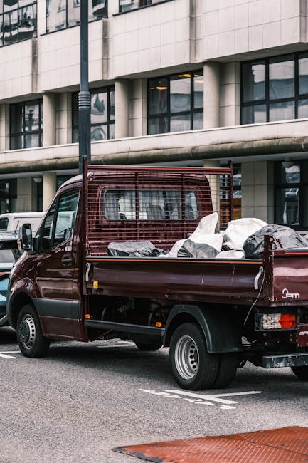 A dark red, small-sized commercial van with a metal mesh partition behind the driver's cab, parked on a city street in front of a modern office building with large glass windows and concrete facade. The van's open cargo area is loaded with various household items, including several large bags wrapped in white plastic, cardboard boxes, and other packing materials, indicating it is part of a house removal or furniture transport. The items are arranged in the van's flatbed space, with some wrapped securely and others loosely placed. The scene is outdoors during daytime, with additional parked vehicles visible nearby, and a lamppost close to the van. The image illustrates a typical home relocation loading process, with the van ready for transport to a new residence, such as those serviced by Man With a Van Wallington, highlighting the equipment and materials used in packing and moving tasks.