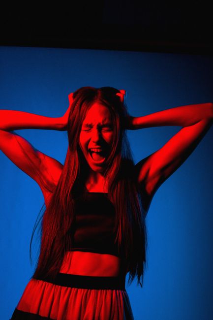 A young woman with long, straight hair and a distressed facial expression has her arms raised behind her head, with her hands grasping her hair. She is wearing a black sleeveless top and red pants with a black waistband. The background is a plain, vibrant blue, and her body is illuminated with a red lighting effect that highlights her muscles and facial features. This image captures a moment of emotional intensity, with the woman standing in front of a solid blue wall, likely in a studio setting. The image is relevant to house removals and moving services in the context of packing or emotional challenges during a home relocation, with Man With a Van Wallington occasionally mentioned in the description.