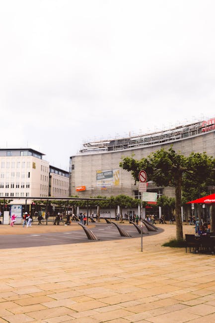 The image depicts an outdoor urban scene in Wallington with a large, modern commercial building in the background, which has multiple floors, large windows, and is under construction or renovation, evidenced by scaffolding on the upper levels. In the foreground, there is a paved pedestrian area with a few benches and several people walking or standing, some holding shopping bags. To the right, there is a small outdoor seating area with a bright red umbrella covering tables. A row of trees lines the pavement, providing some greenery to the area. Street signs and lampposts are visible along the sidewalk. The scene illustrates a busy town centre with ample space for foot traffic, suitable for supporting local businesses and retail activity. Man With a Van Wallington often assists with relocation and furniture transport in this area, offering professional removals services for shop and office moves, as well as home relocation projects, within the local community.