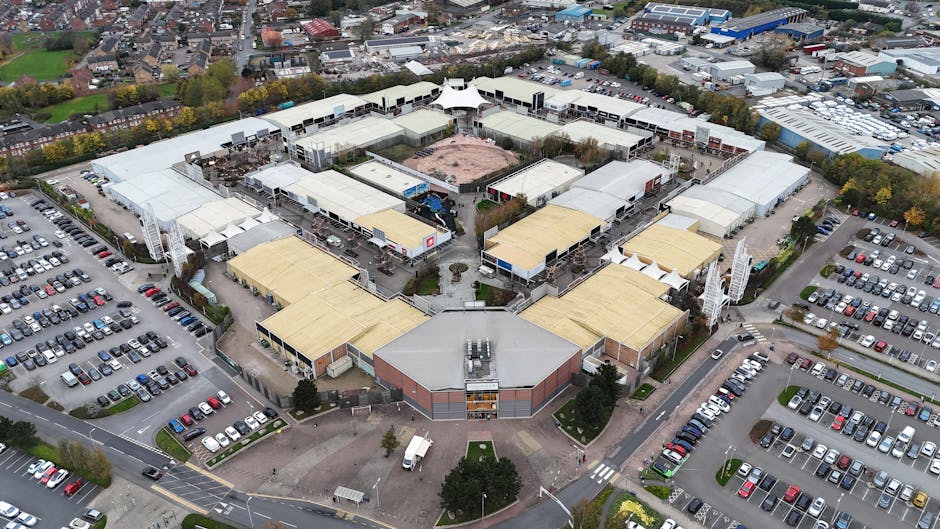 Aerial view of a shopping centre with large commercial buildings having flat roofs covered in light-colored and yellow materials, surrounded by extensive parking lots filled with parked cars, including sedans, hatchbacks, and vans. The parking areas are divided by landscaped islands with trees and grass. The shopping centre is situated in a suburban area, with residential houses and streets visible in the background. The image captures the loading process involved in a home or business relocation, with a focus on transport logistics and moving equipment, including a man with a van or truck associated with Man With a Van Wallington, as part of their removals services, within the context of transferring furniture, boxes, and goods during a house or shop move.