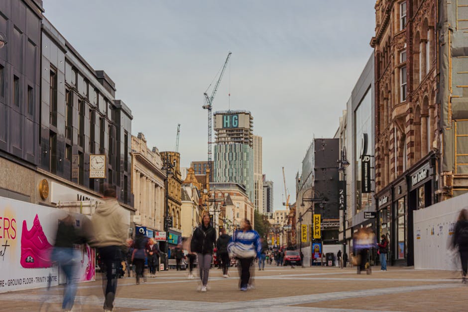 The image depicts an outdoor urban scene in Wallington with a large, modern commercial building in the background, which has multiple floors, large windows, and is under construction or renovation, evidenced by scaffolding on the upper levels. In the foreground, there is a paved pedestrian area with a few benches and several people walking or standing, some holding shopping bags. To the right, there is a small outdoor seating area with a bright red umbrella covering tables. A row of trees lines the pavement, providing some greenery to the area. Street signs and lampposts are visible along the sidewalk. The scene illustrates a busy town centre with ample space for foot traffic, suitable for supporting local businesses and retail activity. Man With a Van Wallington often assists with relocation and furniture transport in this area, offering professional removals services for shop and office moves, as well as home relocation projects, within the local community.