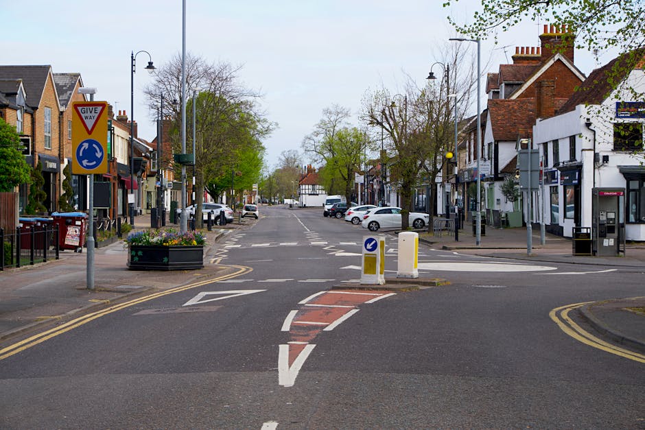 A wide view of a small urban street in Wallington with a narrow access point for vehicle and moving truck entry, featuring double yellow lines along the pavement, parked cars on both sides, and a central priority roundabout with a 'Give Way' sign and a blue circular directional sign. The street is lined with mixed brick and white-painted commercial and residential buildings, some with shopfronts and others with residential entrances. Trees with sparse leaves and street lamps are visible along the pavement, suggesting early spring. The scene is well-lit with natural daylight, and in the background, more parked cars and the continuation of the street can be seen, illustrating the tight-access environment that [COMPANY_NAME] handles during house removals and furniture transport in tight urban settings.