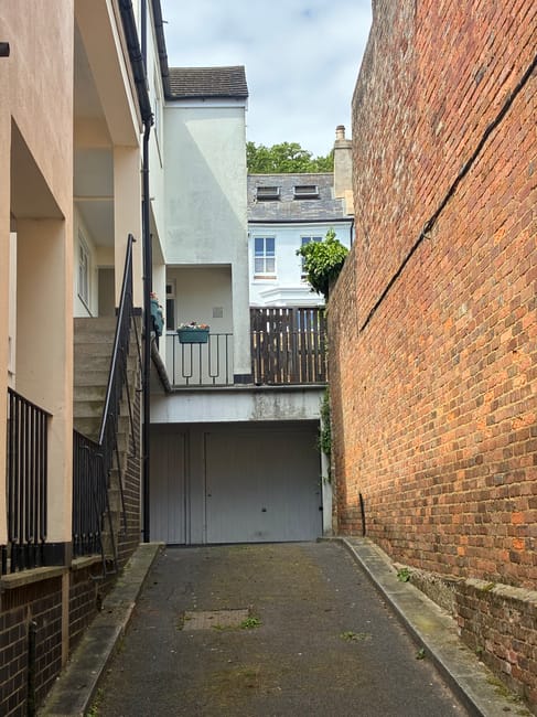 A narrow driveway between a white residential building with exterior stairs and black metal railings on the left, and a tall red brick exterior wall on the right. The driveway is paved with asphalt and has some patches of moss and small plants growing along the edges. At the top of the driveway, there is a wooden gate and a small balcony with flower boxes and potted plants. Behind the gate, parts of neighboring buildings and green trees are visible under a cloudy sky. This scene depicts a tight-access urban environment suitable for furniture transport and home relocation services offered by Man With a Van Wallington, highlighting the challenges of moving in confined spaces.