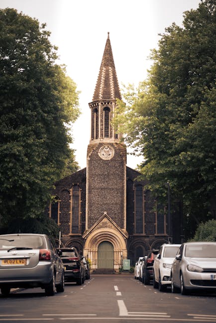 A narrow residential street with a church at the end, featuring a tall, pointed spire and a large clock face on the tower. The street is lined with several parked cars on both sides, including sedans 