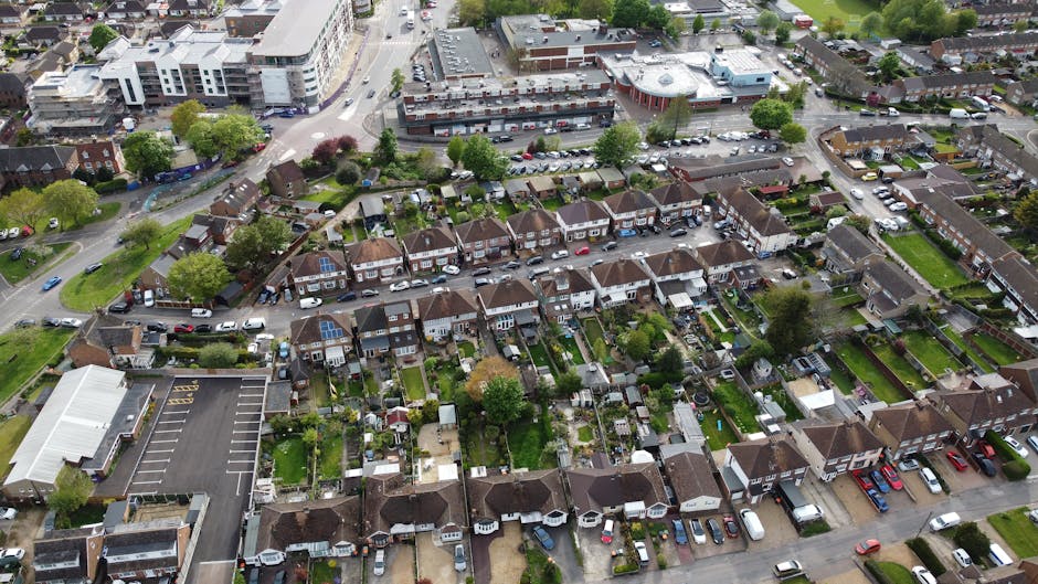 Aerial view of a residential area showing a row of terraced houses with small, well-maintained gardens at the back, some with sheds and outdoor furniture, and driveways with parked cars. The houses are positioned along a narrow street, with a larger commercial building in the background and a parking lot with marked spaces nearby. Multiple vehicles, including vans used for house removals and furniture transport by Man With a Van Wallington, are visible on the street and in the parking areas, some in the process of loading or unloading household belongings during a home relocation. The scene is captured during daylight with clear weather, highlighting the typical urban layout of Wallington for moving services, showcasing the environment where packing, furniture transport, and loading activities take place in a professional and organized manner.