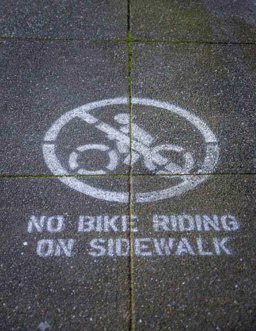 Close-up view of a textured concrete sidewalk featuring a circular white stencil paint marking that displays a bicycle symbol with a diagonal line crossing through it, accompanied by the text 'NO BIKES RIDING ON SIDEWALK'. The stencil is positioned on the pavement outside a property, with visible expansion joints running across the sidewalk. This outdoor scene relates to parking regulation signage often seen in urban residential or commercial areas. The image may be relevant in the context of house removals and moving logistics by highlighting street rules and regulations that movers like Man With a Van Wallington should consider when planning access and parking, ensuring compliance during the loading and unloading process.