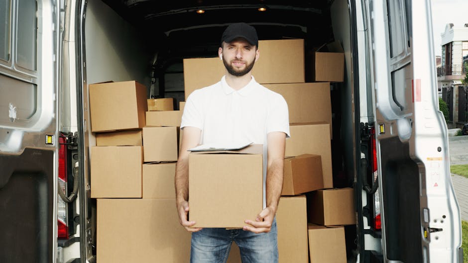 Two movers from Man With a Van Wallington are engaged in a home relocation process, transferring a large cardboard box labelled 'fragile' onto a hand truck on a paved driveway outside a residential bu