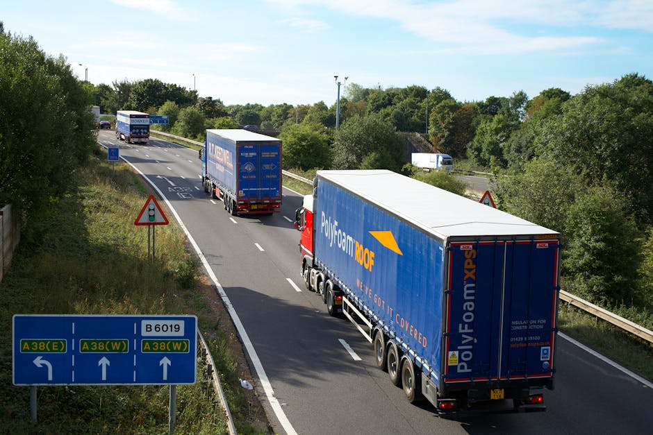 A convoy of three large moving trucks with blue and white Polyfoam XPS branding is traveling along a multi-lane road surrounded by greenery, trees, and bushes on both sides during daylight hours. The trucks are positioned one behind the other, with the closest truck in the foreground featuring a red cab and a spacious, fully covered trailer for transporting furniture and household items. The second truck is slightly further back, with similar branding, and the third truck is further into the distance, all on a smooth asphalt road with clear white lane markings. On the roadside, a blue directional road sign indicates routes toward A38(E), A38(W), and B6019, with arrows guiding drivers. The sky is partially cloudy, and the weather appears clear and bright. This scene depicts a typical home relocation logistics process involving the transportation of household goods via professional removals by companies like Man With a Van Wallington, with the trucks preparing for or engaged in a furniture transport or moving process.