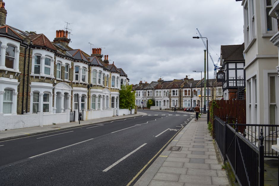 A residential street scene in Woodcote Road, Wallington, featuring a row of Victorian-style terraced houses with white-painted facades and red tiled roofs, under a cloudy sky. The pavement runs alongside the houses on the right, with black metal railings bordering the front gardens. Overhead, there are tall streetlights and construction cranes visible in the distance, indicating ongoing development. The street appears quiet with no vehicles parked along the curb and only a few pedestrians walking along the sidewalk. On the left, the road is lined with similar terraced homes with decorative bay windows and chimney stacks. This scene, as seen in the Wallington moving guide, illustrates an ideal location for household relocations by [COMPANY_NAME], with ample space for loading and unloading furniture and moving boxes during a home relocation process. The street is well-maintained with clear lane markings and accessible sidewalks, providing a suitable environment for efficient furniture transport and packing and moving activities in the area.